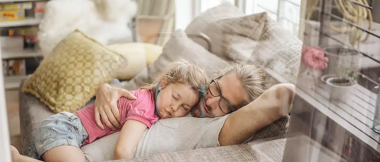 Ein junger Mann mit seiner Tochter schläft bequem auf dem Sofa. Sie sind dank Schallschutzfenstern bestens vor Lärm geschützt.