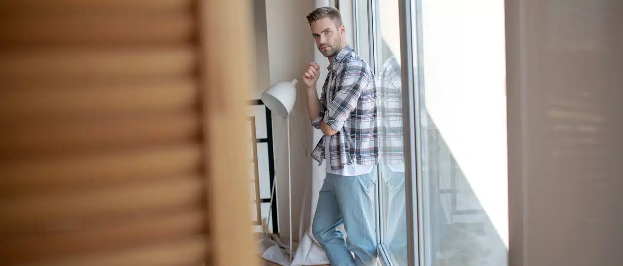 Richtige Größe der Balkontüre finden - Moderner junger Mann steht in heller Wohnung vor großen Kunststoff-Balkontüren.