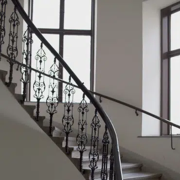 Ein altes Treppenhaus in einem Altbau mit einem Stiegenhausfenster im Hintergrund und einem Schmiedeeisengelände.